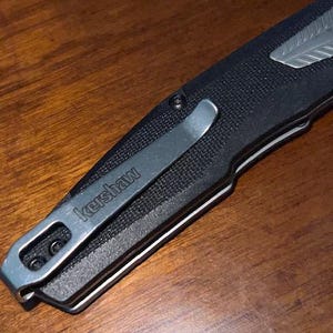 May include: Close-up of a black folding knife with a silver pocket clip. The clip is engraved with the word "kershaw". The knife has a textured handle and a silver accent near the blade. The knife is resting on a wooden surface.