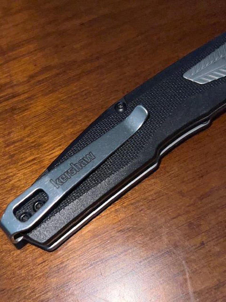 May include: Close-up of a black folding knife with a silver pocket clip. The clip is engraved with the word "kershaw". The knife has a textured handle and a silver accent near the blade. The knife is resting on a wooden surface.