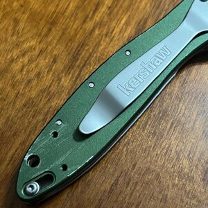 May include: Close-up of a green folding knife with a silver pocket clip. The word "Kershaw" is printed in silver on the clip. The knife is on a wooden surface.