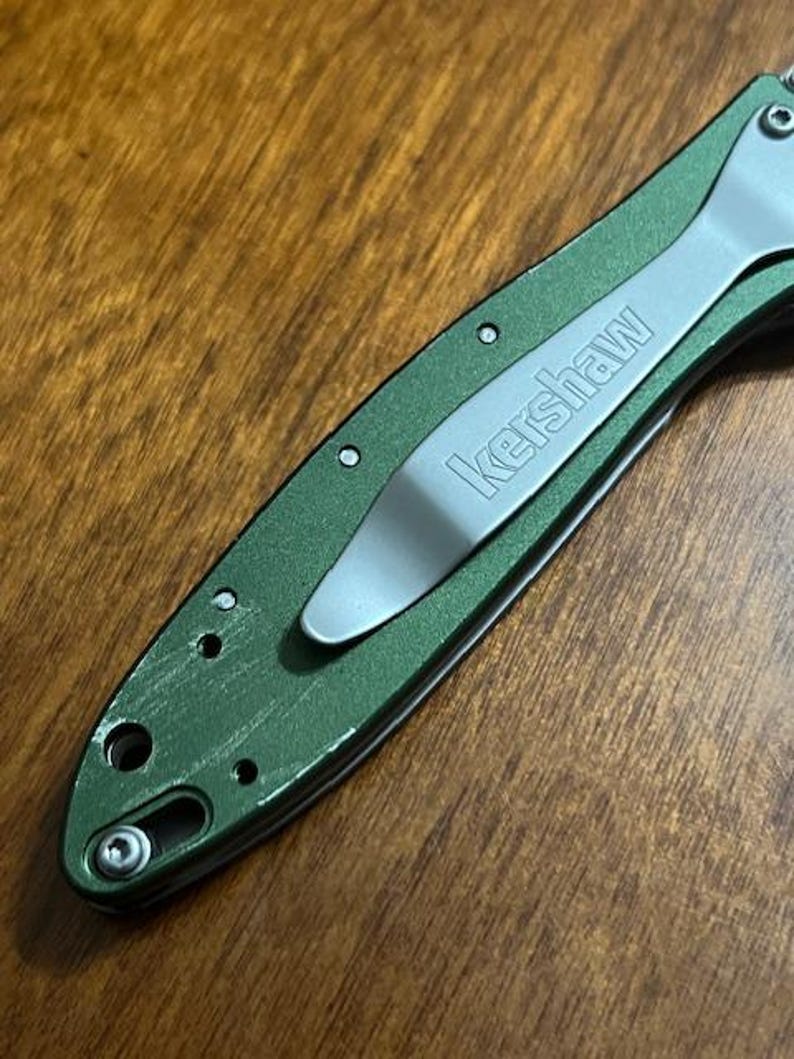 May include: Close-up of a green folding knife with a silver pocket clip. The word "Kershaw" is printed in silver on the clip. The knife is on a wooden surface.