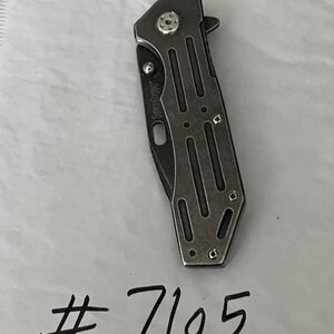 Puede incluir: Una navaja de bolsillo plegable Kershaw cerrada con un mango gris met&aacute;lico que presenta recortes verticales y un tornillo de pivote visible. La navaja est&aacute; colocada sobre el texto escrito a mano # 7105 en una superficie blanca texturizada.