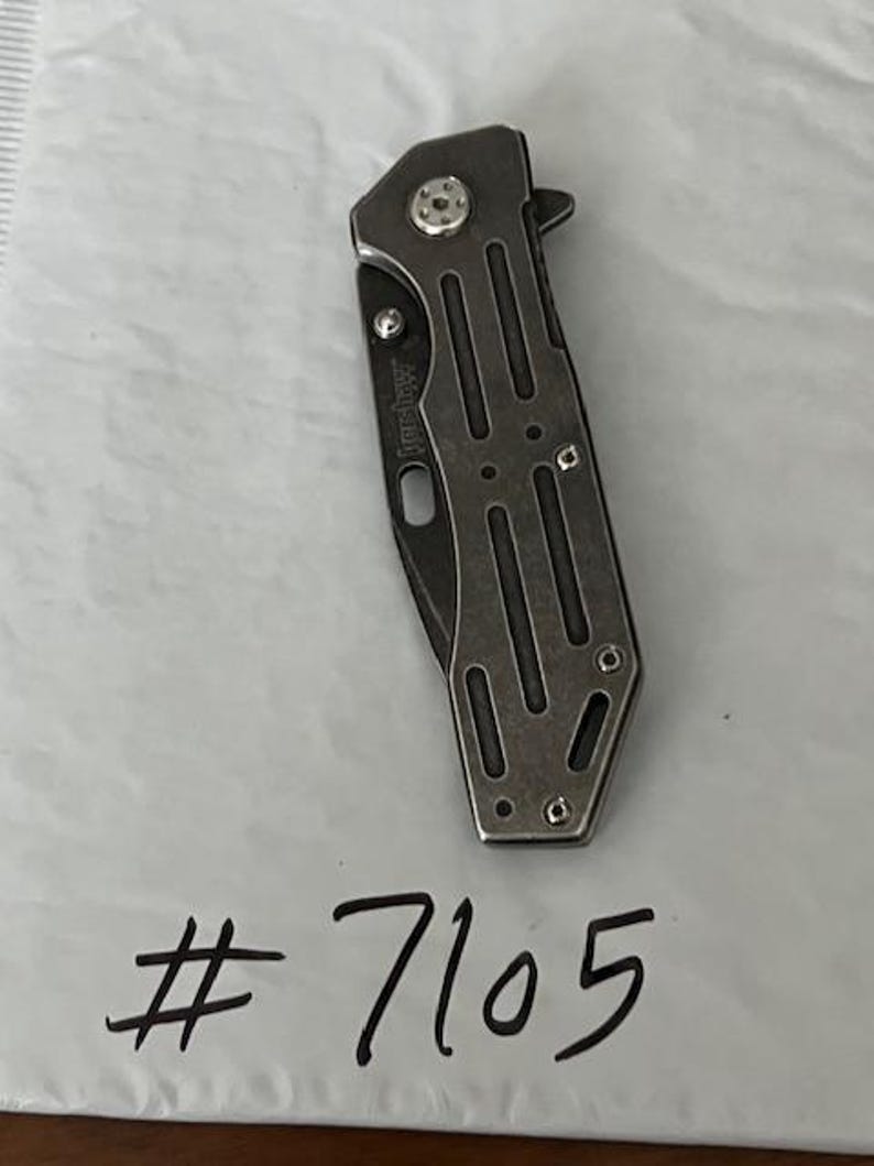 Puede incluir: Una navaja de bolsillo plegable Kershaw cerrada con un mango gris met&aacute;lico que presenta recortes verticales y un tornillo de pivote visible. La navaja est&aacute; colocada sobre el texto escrito a mano # 7105 en una superficie blanca texturizada.