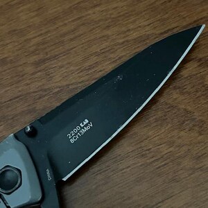 K&ouml;nnte beinhalten: Ein schwarzes Klappmesser mit einer teilweise gezahnten Klinge. Die Klinge tr&auml;gt den Text "2200 KAI 8Cr13MoV" und der Griff ist grau. Das Messer liegt auf einer Holzoberfl&auml;che.