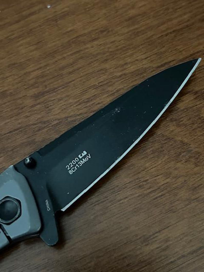 K&ouml;nnte beinhalten: Ein schwarzes Klappmesser mit einer teilweise gezahnten Klinge. Die Klinge tr&auml;gt den Text "2200 KAI 8Cr13MoV" und der Griff ist grau. Das Messer liegt auf einer Holzoberfl&auml;che.