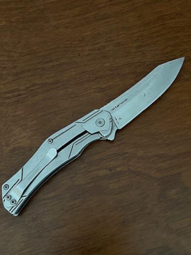 May include: A silver folding knife with a partially serrated blade. The knife has a clip for pocket carry and a textured handle. The blade is marked with the text "1340 EA Patented".
