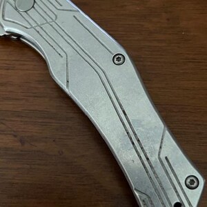 May include: Close-up of a silver folding knife with a textured handle. The knife blade is partially visible, and the handle features a series of parallel lines and geometric shapes. The knife is on a dark brown wooden surface.
