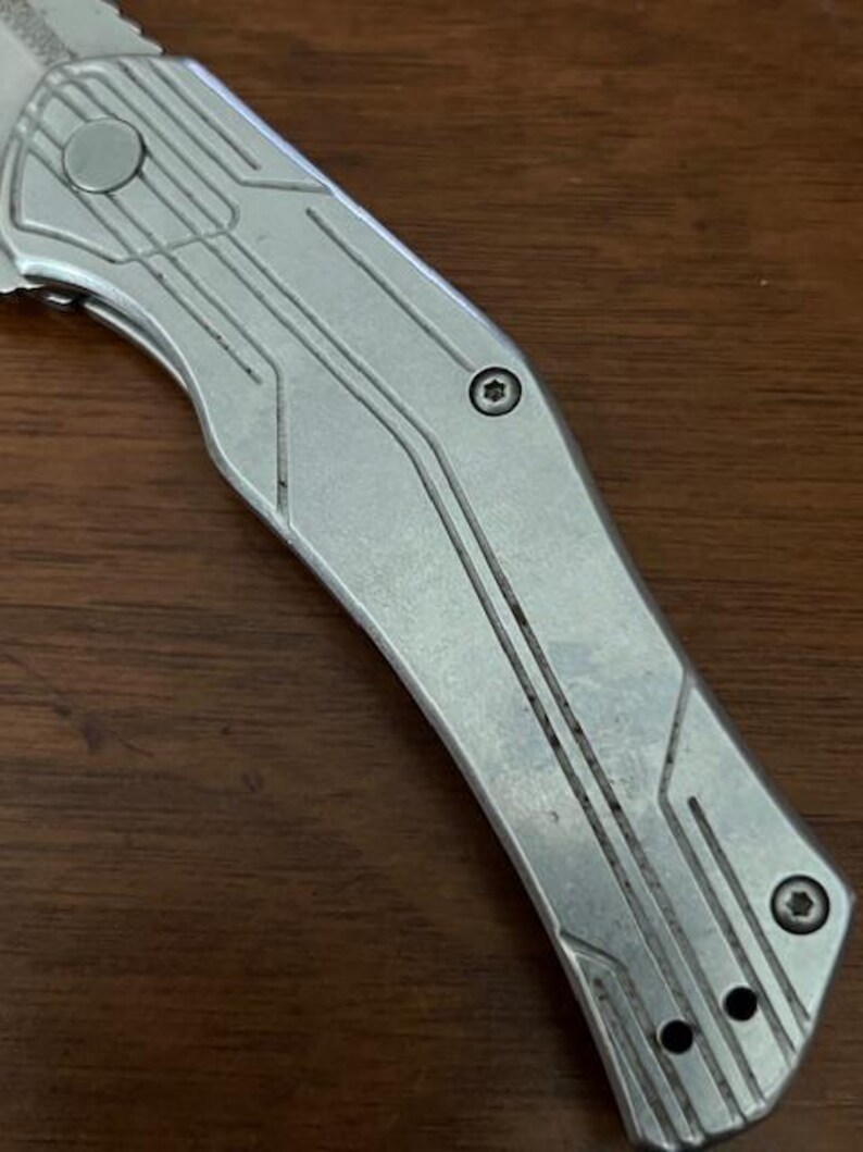 May include: Close-up of a silver folding knife with a textured handle. The knife blade is partially visible, and the handle features a series of parallel lines and geometric shapes. The knife is on a dark brown wooden surface.