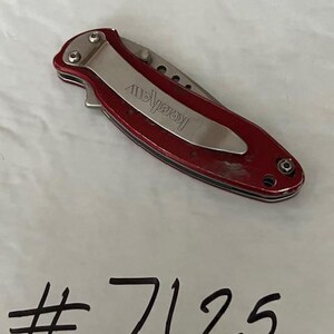 May include: A closed folding pocket knife with a metallic red handle and a silver pocket clip featuring the brand name Kershaw engraved on the clip. Below the knife, the number #7125 is written in black marker on the white surface.