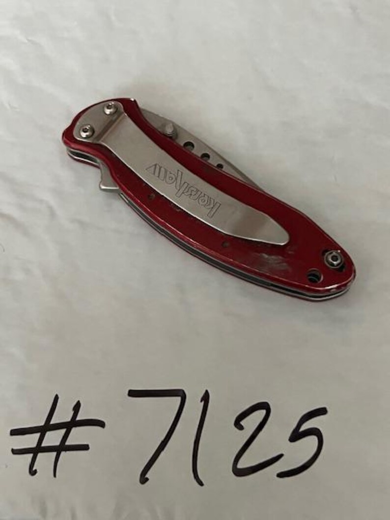 May include: A closed folding pocket knife with a metallic red handle and a silver pocket clip featuring the brand name Kershaw engraved on the clip. Below the knife, the number #7125 is written in black marker on the white surface.
