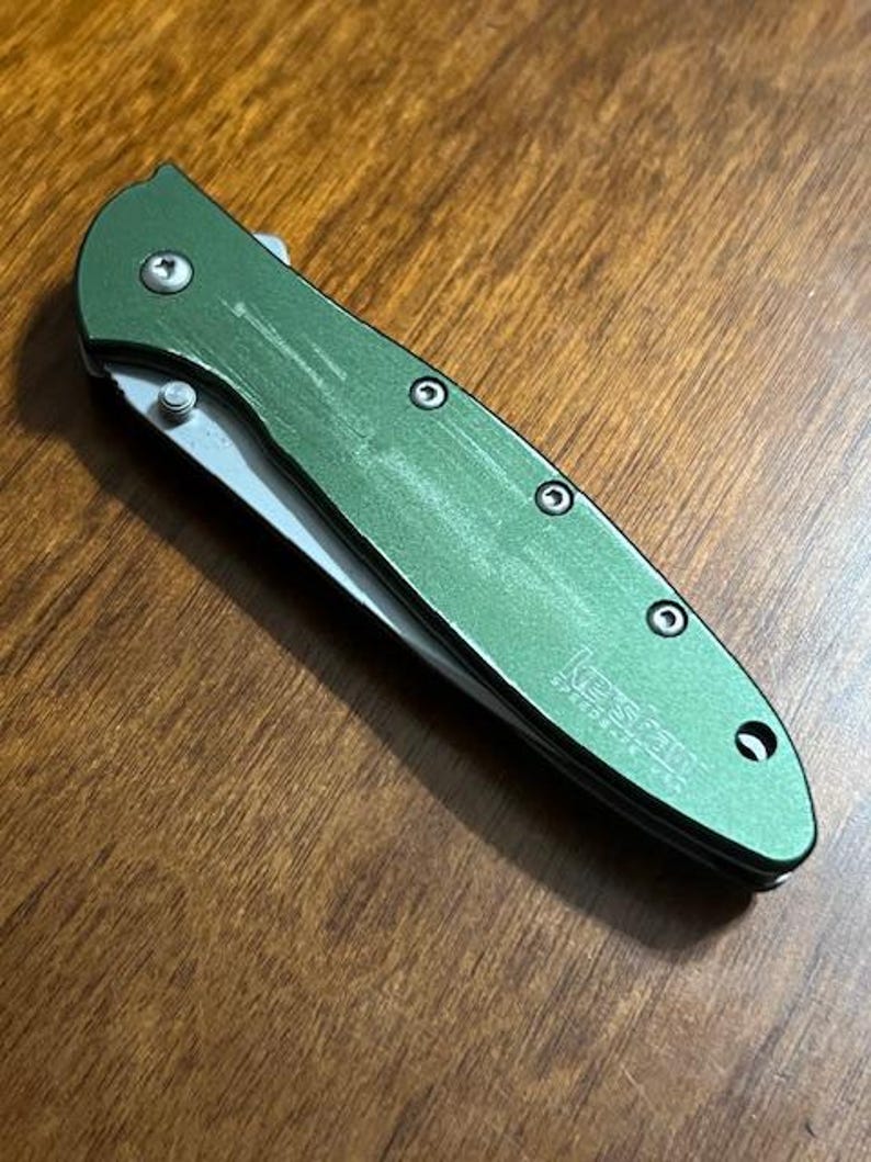 May include: A green folding pocket knife with a silver blade and a textured handle. The knife has the brand name "Kershaw" printed on the handle. The knife is lying on a wooden surface.