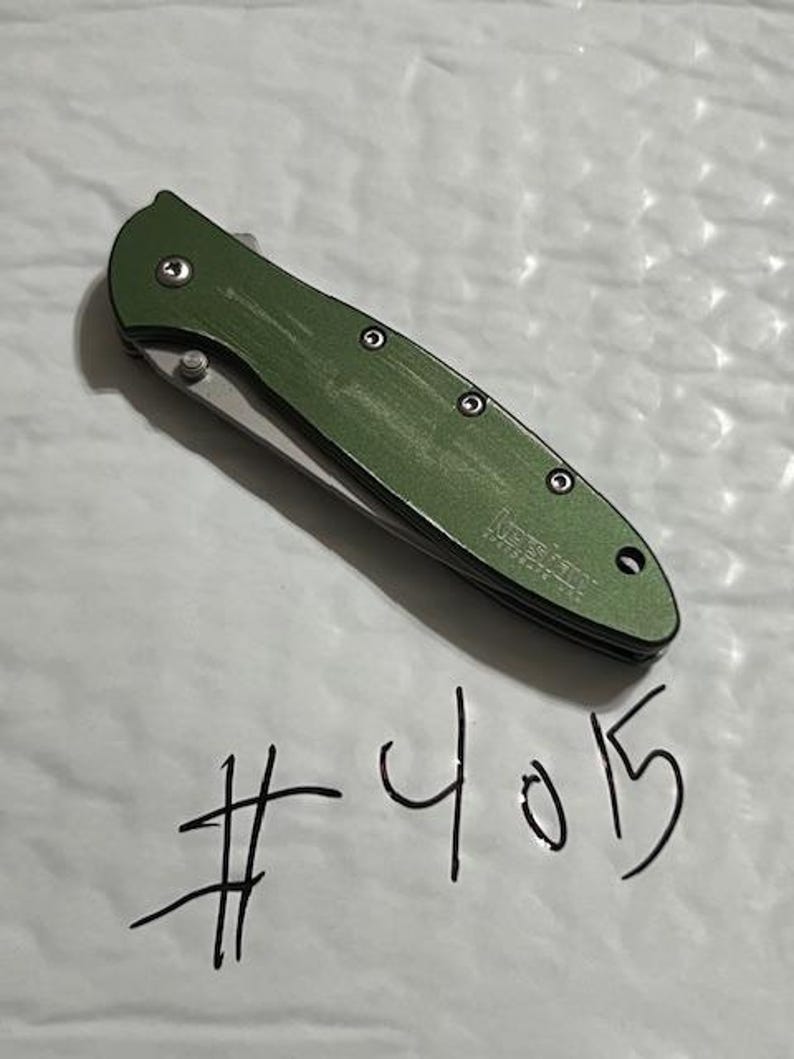 May include: A green folding pocket knife with a silver blade. The knife has a textured handle and is labeled "Kershaw". The number "4015" is handwritten below the knife.