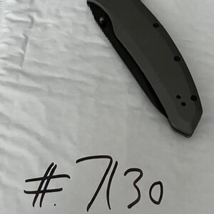 K&ouml;nnte beinhalten: Ein dunkelgraues Klappmesser mit schwarzer Klinge liegt auf einer wei&szlig;en Oberfl&auml;che. Das Messer hat einen strukturierten Griff und einen runden Knopf. Die handgeschriebene Nummer "7130" in schwarzer Tinte befindet sich darunter.