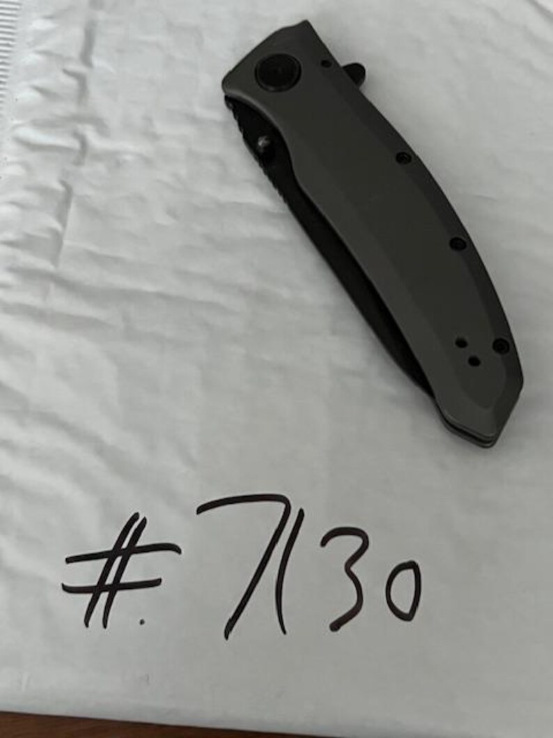 K&ouml;nnte beinhalten: Ein dunkelgraues Klappmesser mit schwarzer Klinge liegt auf einer wei&szlig;en Oberfl&auml;che. Das Messer hat einen strukturierten Griff und einen runden Knopf. Die handgeschriebene Nummer "7130" in schwarzer Tinte befindet sich darunter.