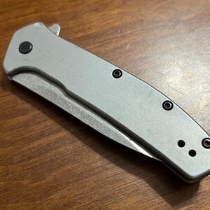 May include: A silver folding knife with a partially exposed blade. The knife has a textured handle with black screws and a black button. The blade is silver and the knife is on a wooden surface.