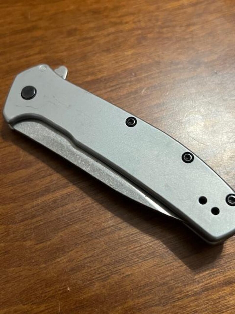 May include: A silver folding knife with a partially exposed blade. The knife has a textured handle with black screws and a black button. The blade is silver and the knife is on a wooden surface.