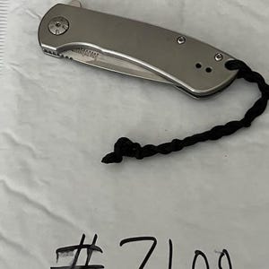 Op de afbeelding: Een zilverkleurig inklapbaar zakmes met een gladde metalen handgreep en een zwart gevlochten koord aan het uiteinde. Het lemmet is gedeeltelijk zichtbaar en bevat de merknaam Kershaw. Het nummer #7100 staat eronder in zwarte inkt geschreven.