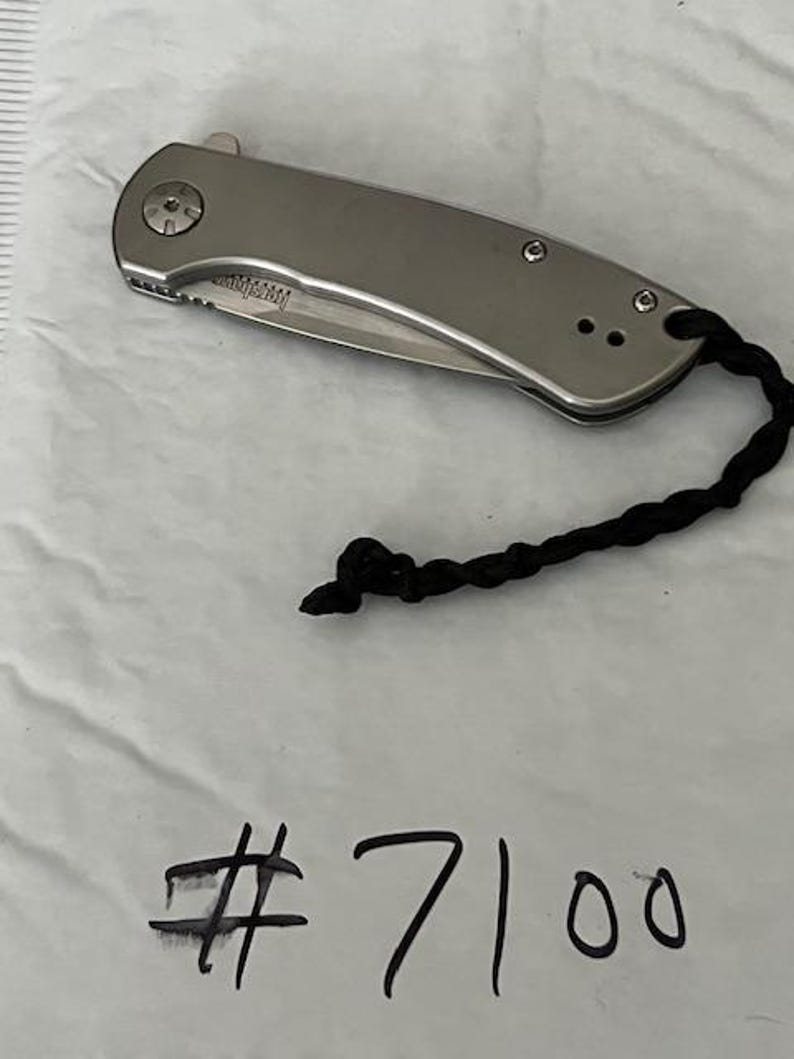 Op de afbeelding: Een zilverkleurig inklapbaar zakmes met een gladde metalen handgreep en een zwart gevlochten koord aan het uiteinde. Het lemmet is gedeeltelijk zichtbaar en bevat de merknaam Kershaw. Het nummer #7100 staat eronder in zwarte inkt geschreven.