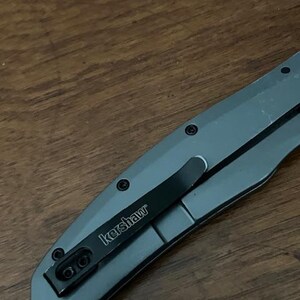 K&ouml;nnte beinhalten: Ein graues Klappmesser mit einem schwarzen Taschenclip. Der Clip ist mit "kershaw" in wei&szlig;er Schrift beschriftet. Das Messer liegt auf einer Holzoberfl&auml;che.