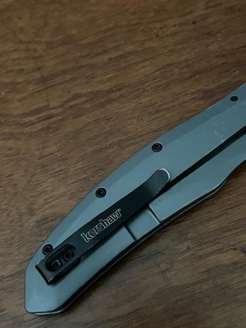 K&ouml;nnte beinhalten: Ein graues Klappmesser mit einem schwarzen Taschenclip. Der Clip ist mit "kershaw" in wei&szlig;er Schrift beschriftet. Das Messer liegt auf einer Holzoberfl&auml;che.
