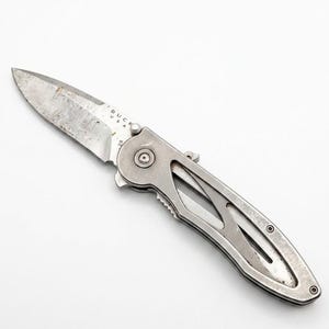 May include: A silver folding knife with a partially serrated blade. The knife has a metal handle with a cutout design and the text "BUCK USA" is visible on the blade. The knife is on a white background.
