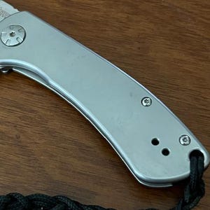 Op de afbeelding: Een zilverkleurig inklapbaar zakmes met een gladde metalen handgreep, voorzien van een opvallende pivotschroef, twee kleinere schroeven en een zwart gevlochten koord dat door een gat aan het uiteinde van de handgreep is bevestigd, met een deel van het lemmet.