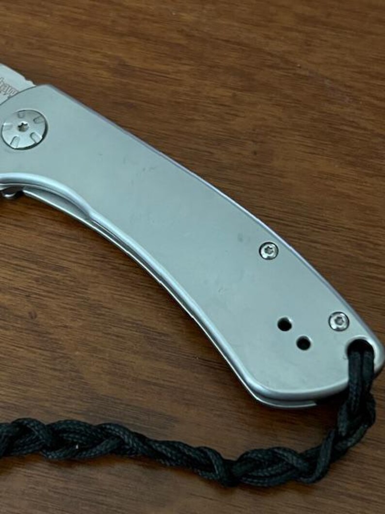 Op de afbeelding: Een zilverkleurig inklapbaar zakmes met een gladde metalen handgreep, voorzien van een opvallende pivotschroef, twee kleinere schroeven en een zwart gevlochten koord dat door een gat aan het uiteinde van de handgreep is bevestigd, met een deel van het lemmet.