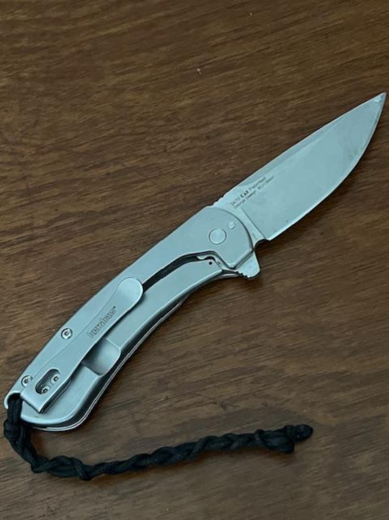 Op de afbeelding: Een zilverkleurig inklapbaar zakmes met een metalen handvat en een scherp lemmet. Op het lemmet staat de tekst 3470 CKT Patented George Design 8Cr13MoV. Aan het uiteinde van het handvat zit een zwart gevlochten koord en een zakclip.