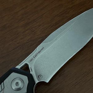 May include: Close-up of a silver and black folding knife. The blade has the text "1390 KAI Patented" etched on it. The handle features a circular design and a black grip.