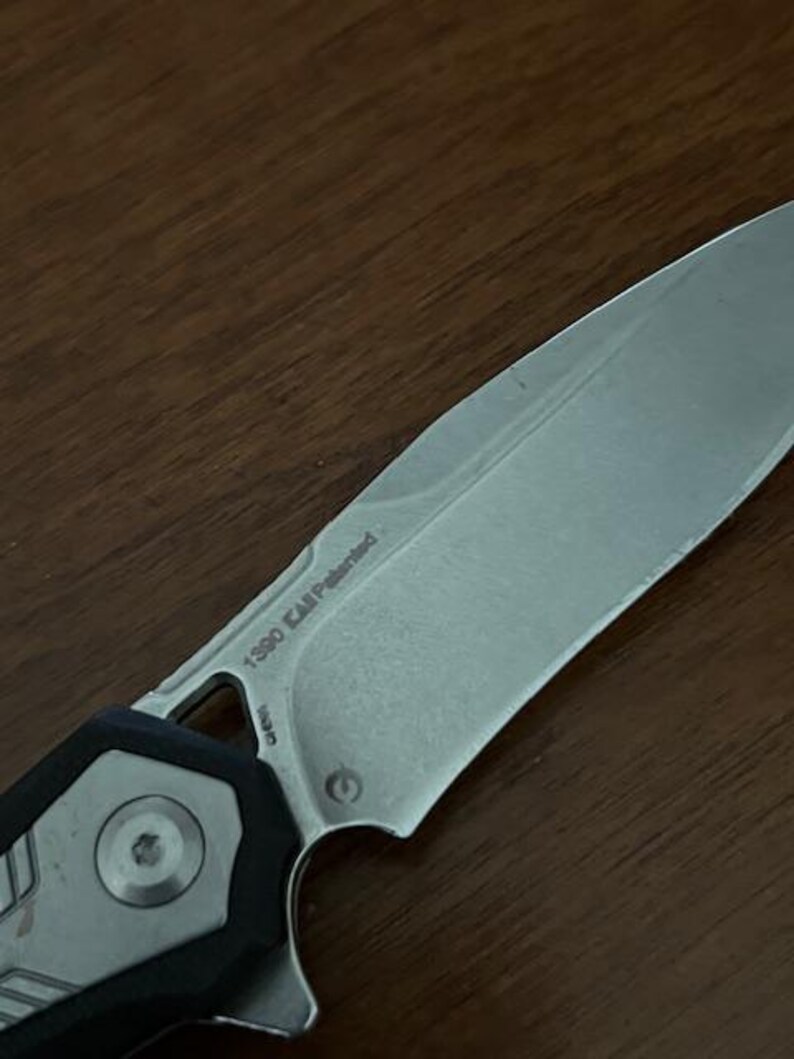 May include: Close-up of a silver and black folding knife. The blade has the text "1390 KAI Patented" etched on it. The handle features a circular design and a black grip.