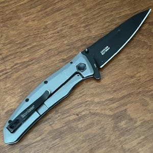 K&ouml;nnte beinhalten: Ein silbernes und schwarzes Klappmesser mit schwarzer Klinge. Das Messer hat einen Taschenclip und den Markennamen "Kershaw". Auf der Klinge steht "2300 SM, 8Cr13MoV". Das Messer liegt auf einer Holzoberfl&auml;che.