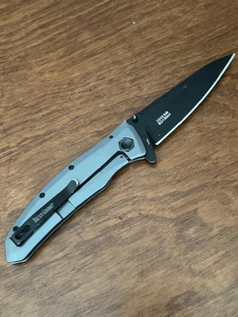 K&ouml;nnte beinhalten: Ein silbernes und schwarzes Klappmesser mit schwarzer Klinge. Das Messer hat einen Taschenclip und den Markennamen "Kershaw". Auf der Klinge steht "2300 SM, 8Cr13MoV". Das Messer liegt auf einer Holzoberfl&auml;che.