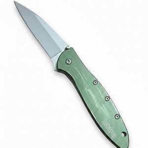 May include: A silver-bladed folding knife with a green handle. The knife has a pointed blade and a handle with three visible rivets. The brand name "Kershaw" is printed on the handle.