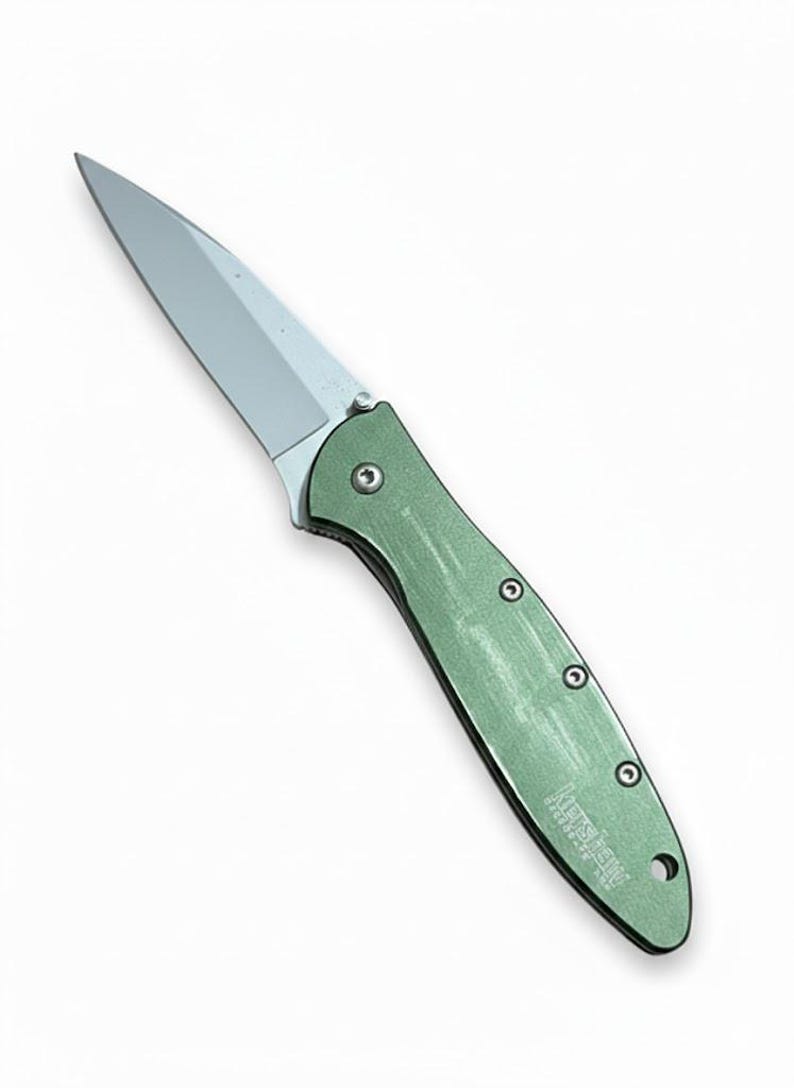 May include: A silver-bladed folding knife with a green handle. The knife has a pointed blade and a handle with three visible rivets. The brand name "Kershaw" is printed on the handle.