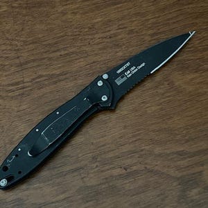 May include: A black folding knife with a partially serrated blade. The knife has a pocket clip and the text "KAI USA" and "Ken Onion Design" is visible. The knife is lying on a wooden surface.