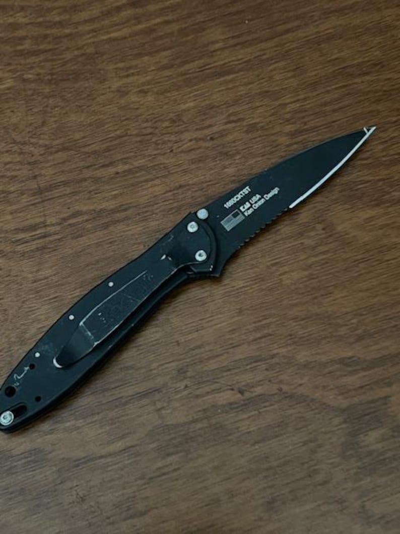 May include: A black folding knife with a partially serrated blade. The knife has a pocket clip and the text "KAI USA" and "Ken Onion Design" is visible. The knife is lying on a wooden surface.