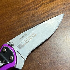 May include: Close-up of a partially open folding knife with a silver blade and a purple handle. The blade has the text "1620PUR" and "KAI Patented Ken Onion Design" printed on it, along with a small American flag graphic.