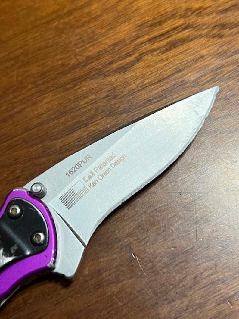 May include: Close-up of a partially open folding knife with a silver blade and a purple handle. The blade has the text "1620PUR" and "KAI Patented Ken Onion Design" printed on it, along with a small American flag graphic.