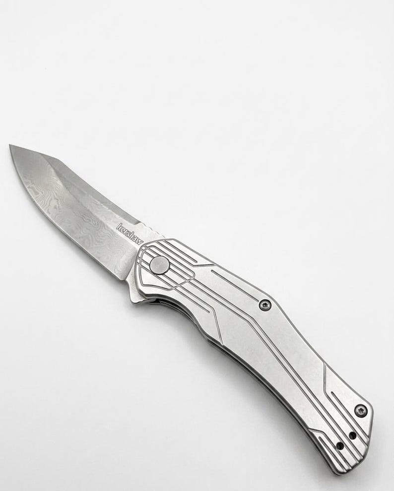 May include: A silver folding knife with a textured blade and handle. The blade has a unique pattern, and the handle features a linear design. The brand name "Kershaw" is visible on the blade.