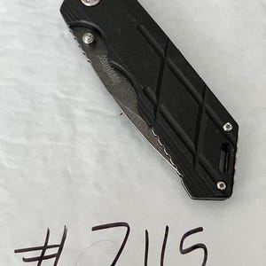 May include: A black folding knife with a partially serrated blade. The knife has a textured handle with a diamond pattern and the brand name "Kershaw" is visible on the blade. The number "7115" is handwritten below the knife.