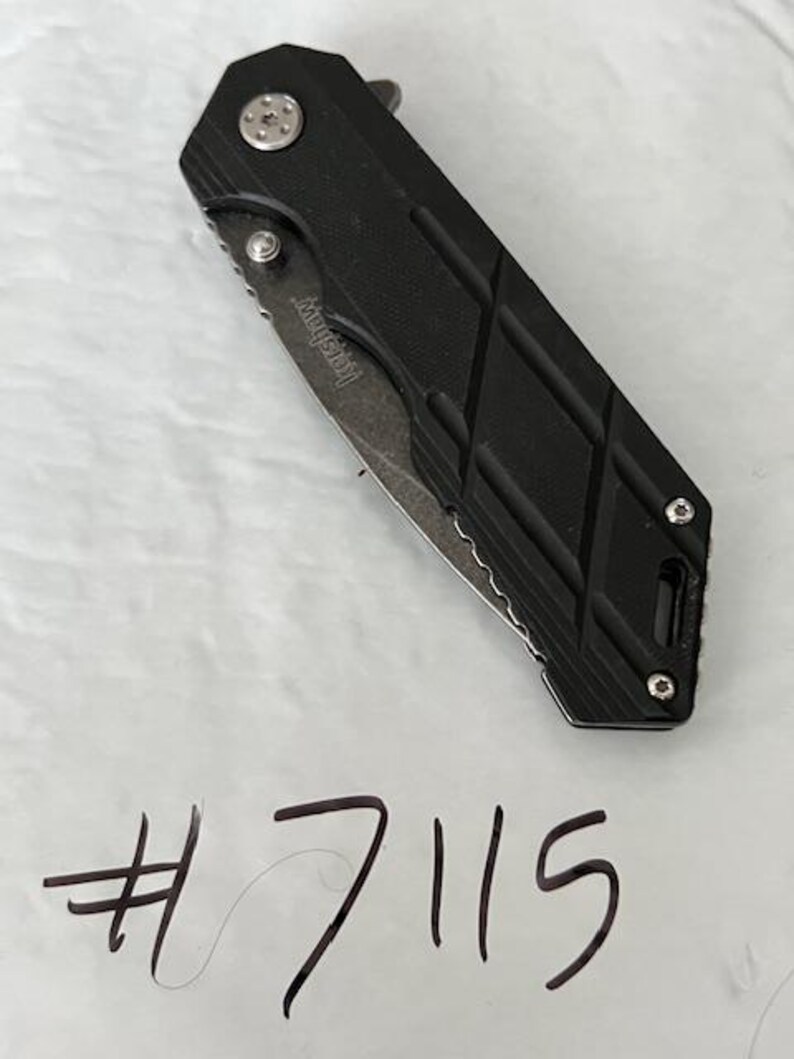 May include: A black folding knife with a partially serrated blade. The knife has a textured handle with a diamond pattern and the brand name "Kershaw" is visible on the blade. The number "7115" is handwritten below the knife.