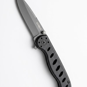 May include: A folding pocket knife with a matte gray blade featuring the word GERBER printed in white. The handle is black with a textured surface and a series of five oval cutouts along the side, secured with visible black screws at the base.