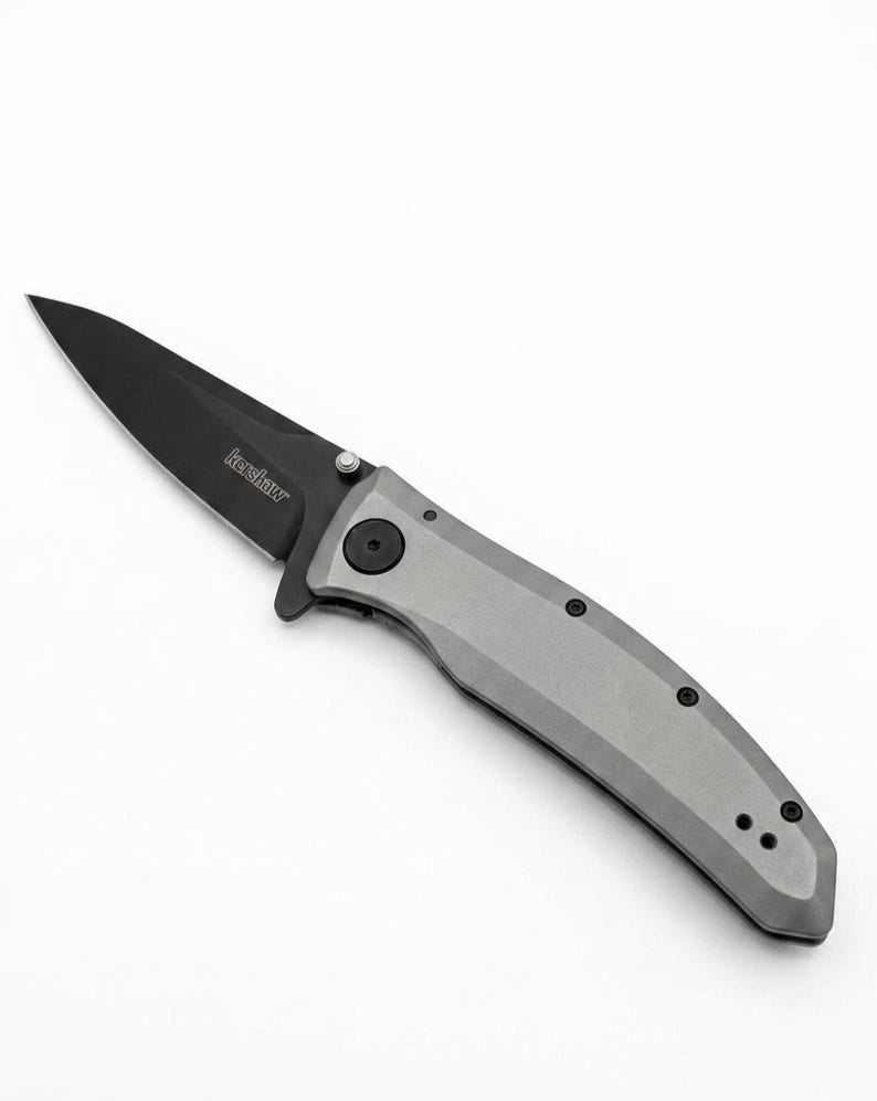K&ouml;nnte beinhalten: Ein Klappmesser mit schwarzer Klinge und grauem Griff. Auf der Klinge ist der Markenname "Kershaw" aufgedruckt. Der Griff hat eine Reihe kleiner schwarzer Schrauben.