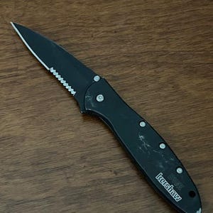May include: Black folding knife with a partially serrated blade. The knife has a black handle with silver rivets and the brand name "Kershaw" printed on the side. The knife is lying on a wooden surface.