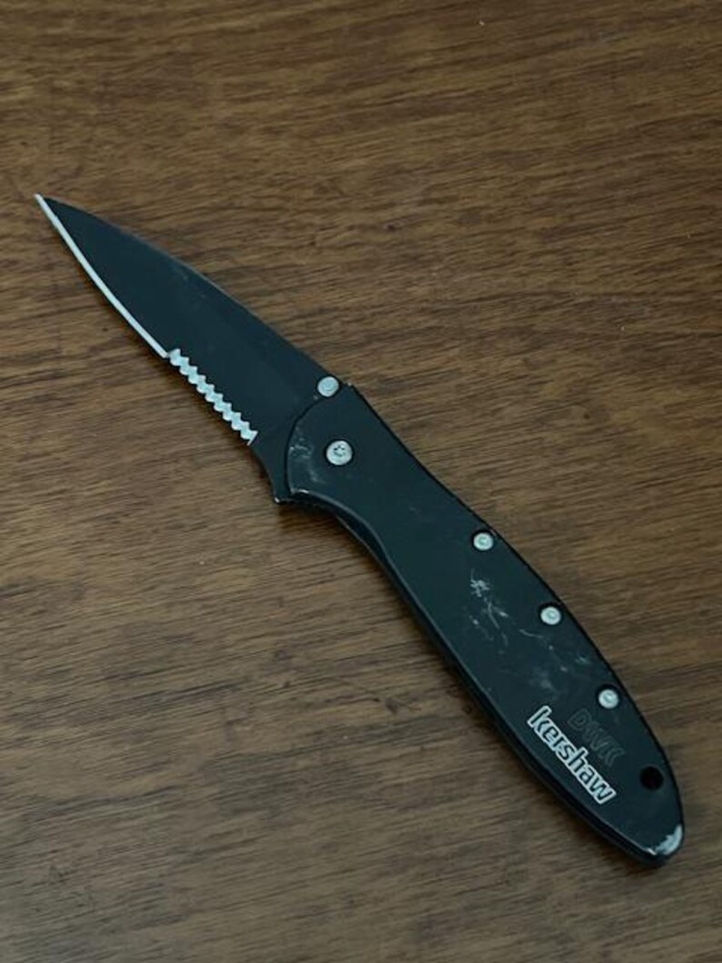 May include: Black folding knife with a partially serrated blade. The knife has a black handle with silver rivets and the brand name "Kershaw" printed on the side. The knife is lying on a wooden surface.