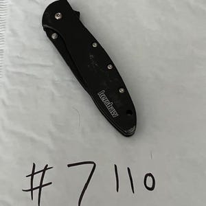May include: A black folding knife with the brand name "Kershaw" visible. The knife is closed and has a series of silver rivets along the handle. The number "7110" is handwritten in black ink below the knife.