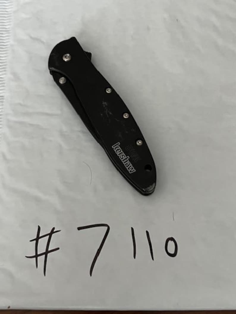 May include: A black folding knife with the brand name "Kershaw" visible. The knife is closed and has a series of silver rivets along the handle. The number "7110" is handwritten in black ink below the knife.