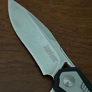 May include: Close-up of a silver and black folding knife. The blade is partially open, revealing the Kershaw Speedsafe logo. The handle is black and gray with a textured design. The knife is on a wooden surface.