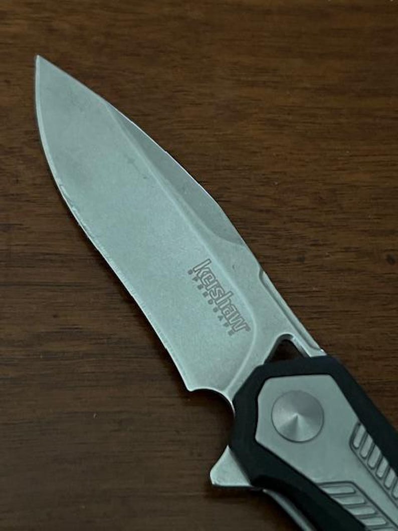 May include: Close-up of a silver and black folding knife. The blade is partially open, revealing the Kershaw Speedsafe logo. The handle is black and gray with a textured design. The knife is on a wooden surface.
