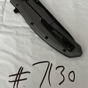 K&ouml;nnte beinhalten: Ein geschlossenes graues Klappmesser mit einem schwarzen Taschenclip mit der Aufschrift "Kershaw". Das Messer hat eine sechseckige Schraube und die Nummer "7130" in schwarzem Marker darunter geschrieben.