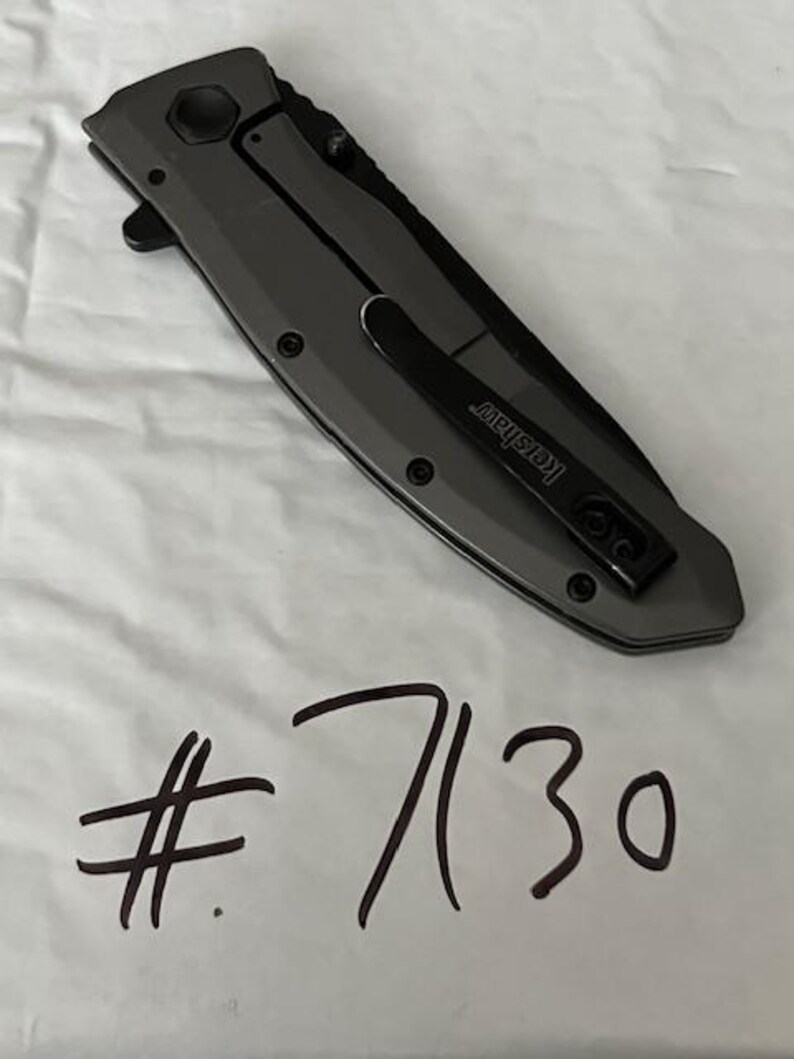 K&ouml;nnte beinhalten: Ein geschlossenes graues Klappmesser mit einem schwarzen Taschenclip mit der Aufschrift "Kershaw". Das Messer hat eine sechseckige Schraube und die Nummer "7130" in schwarzem Marker darunter geschrieben.